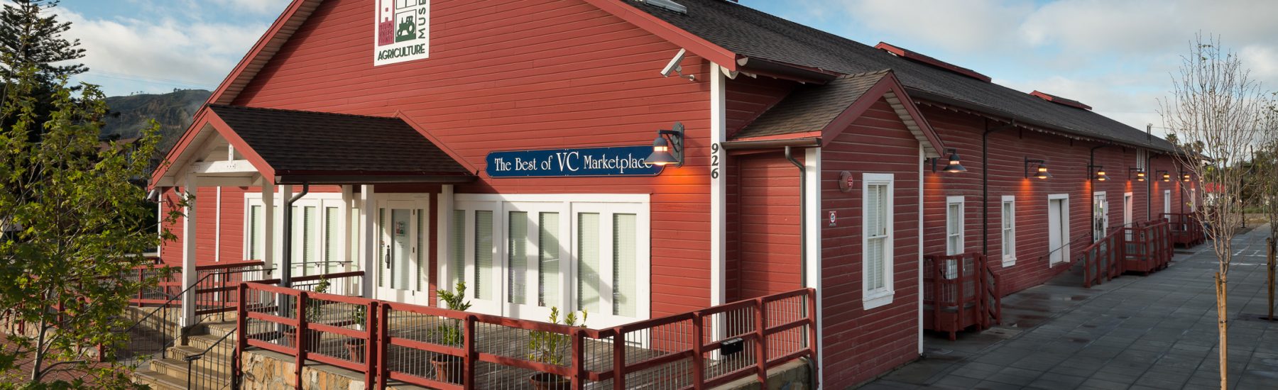 Red building with white trim and stone foundation features "The Best of VC Marketplace" and "Museum Agriculture" signs. Landmark structure from 2013 with railings and sidewalk.