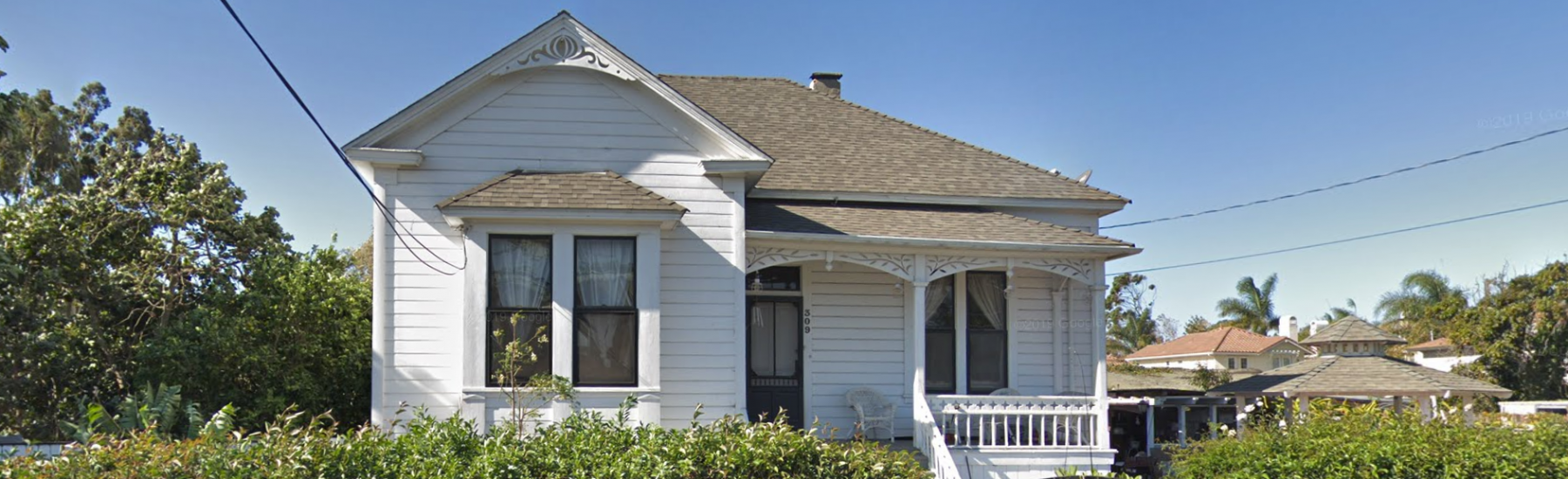 White Victorian house with gray shingled roof and white porch railing, surrounded by dense green hedges. Address number 509 visible on the front door.