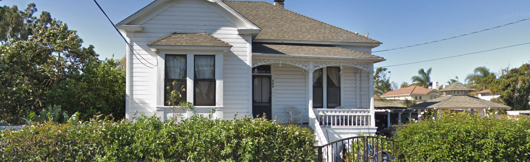 White Victorian house with gray shingled roof and white porch railing, surrounded by dense green hedges. Address number 509 visible on the front door.